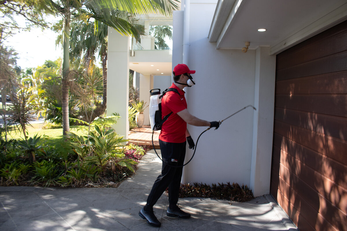 We Kill Termites technician in red uniform spraying pest treatment around home exterior